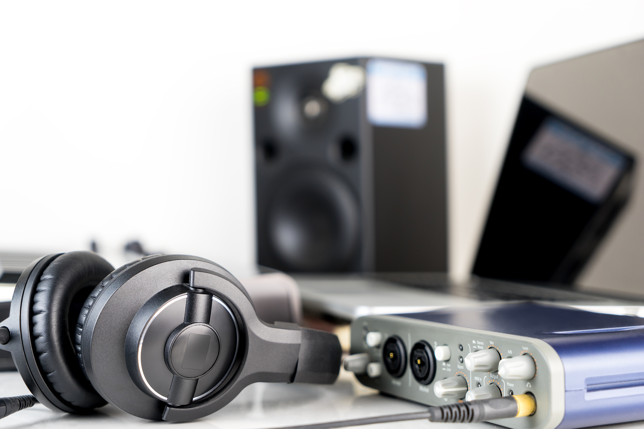Fone de ouvido sobre mesa de trabalho com caixa de som ao fundo e equipamentos de áudio e vídeo ao lado