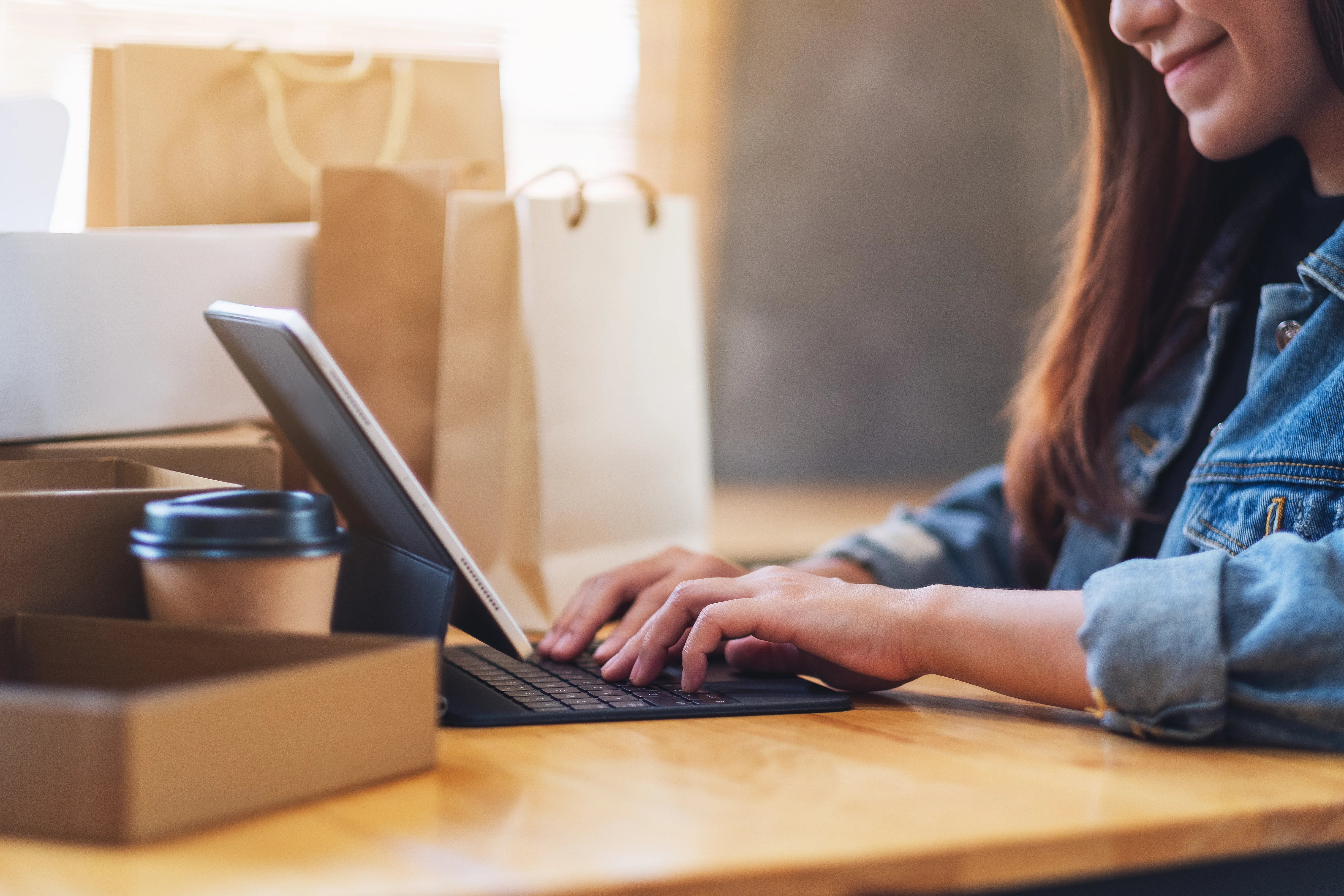 closeup-image-of-a-young-asian-woman-typing-on-tablet-pc-for-online-shopping-with-postal-parcel-box-and-shopping-bags-on-the-table-at-home