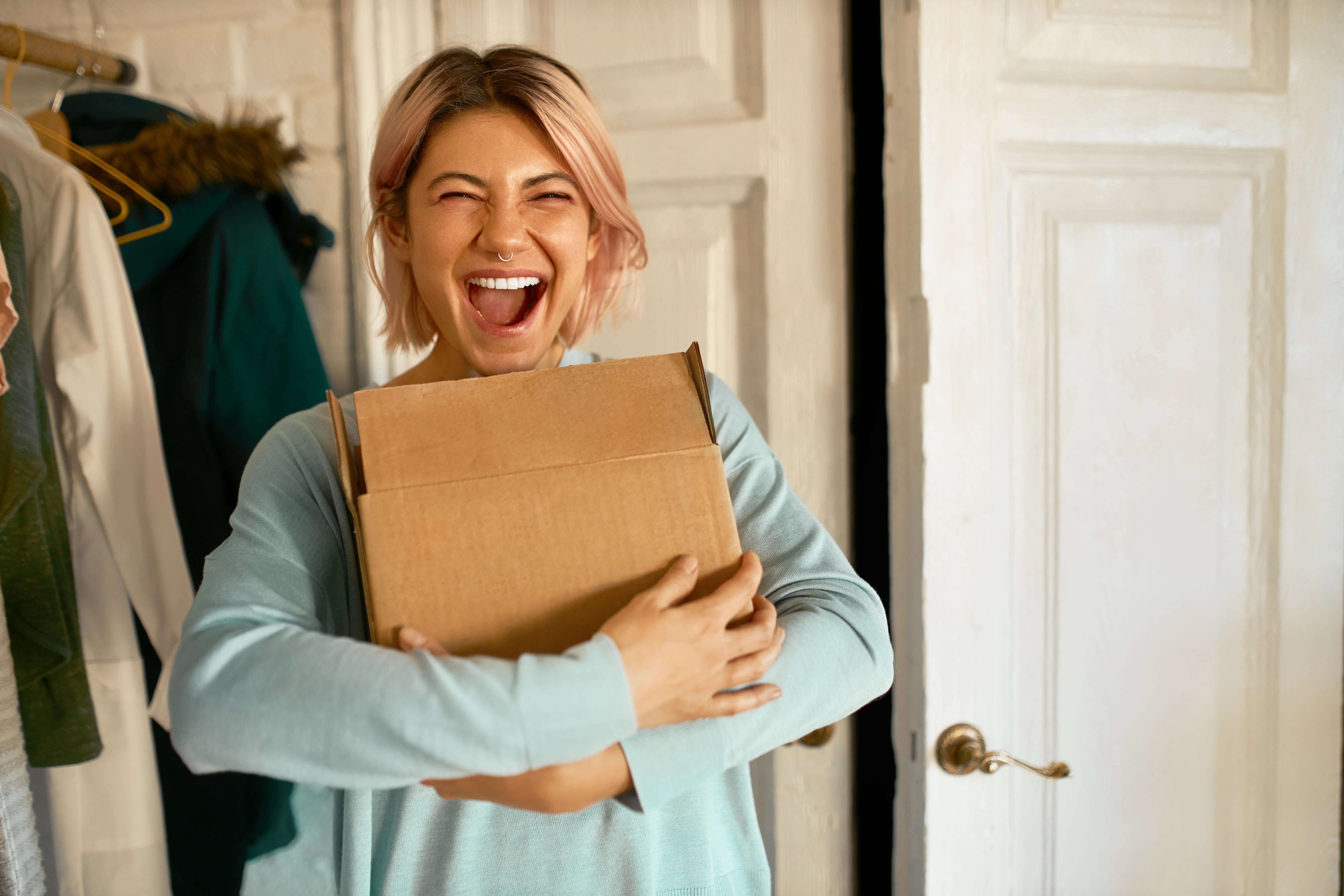 indoor-image-of-happy-cheerful-young-woman-holding-cardboard-box-delivered-to-her-apartment-expressing-excitement-going-to-unpack-parcel