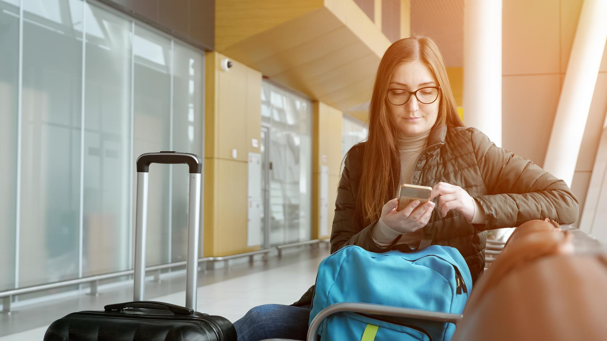young-woman-traveller-passes-e-check-in-mobile-phone-waiting-her-flight-in-airport-she-is-sitting-at-hall-with-her-suitcase-and-backpack-sunlight_Easy-Resize.com